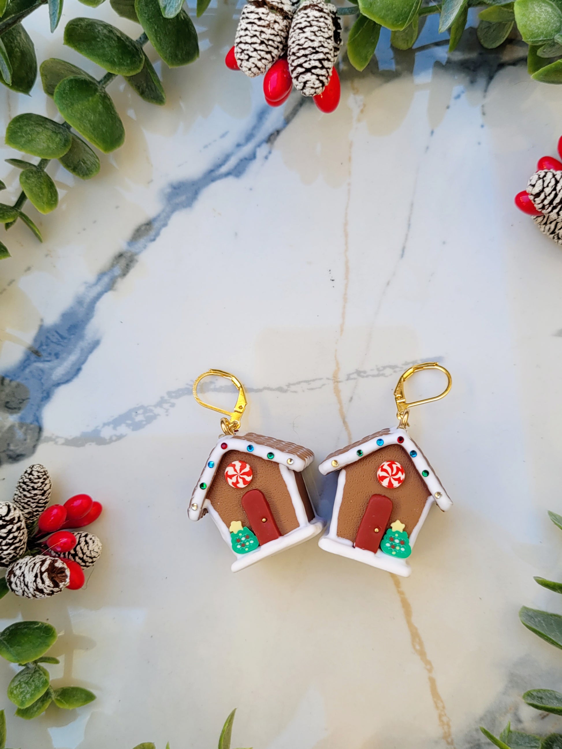 close up of hook Gingerbread house earrings on a marble background surrounded by foliage.
