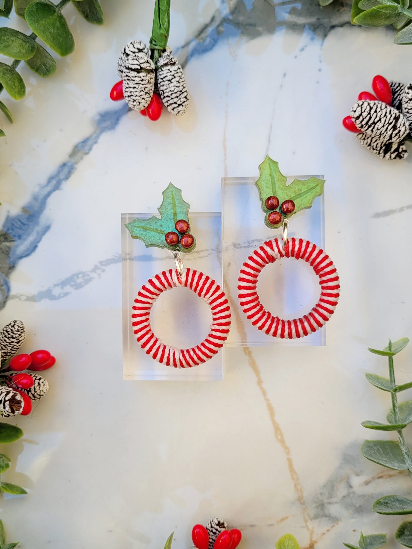 close up of mistletoe and candy cane striped hoop earrings on a white marble background surrounded by foliage.