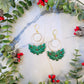 Holly wreath earrings on a marble background surrounded by foliage. 