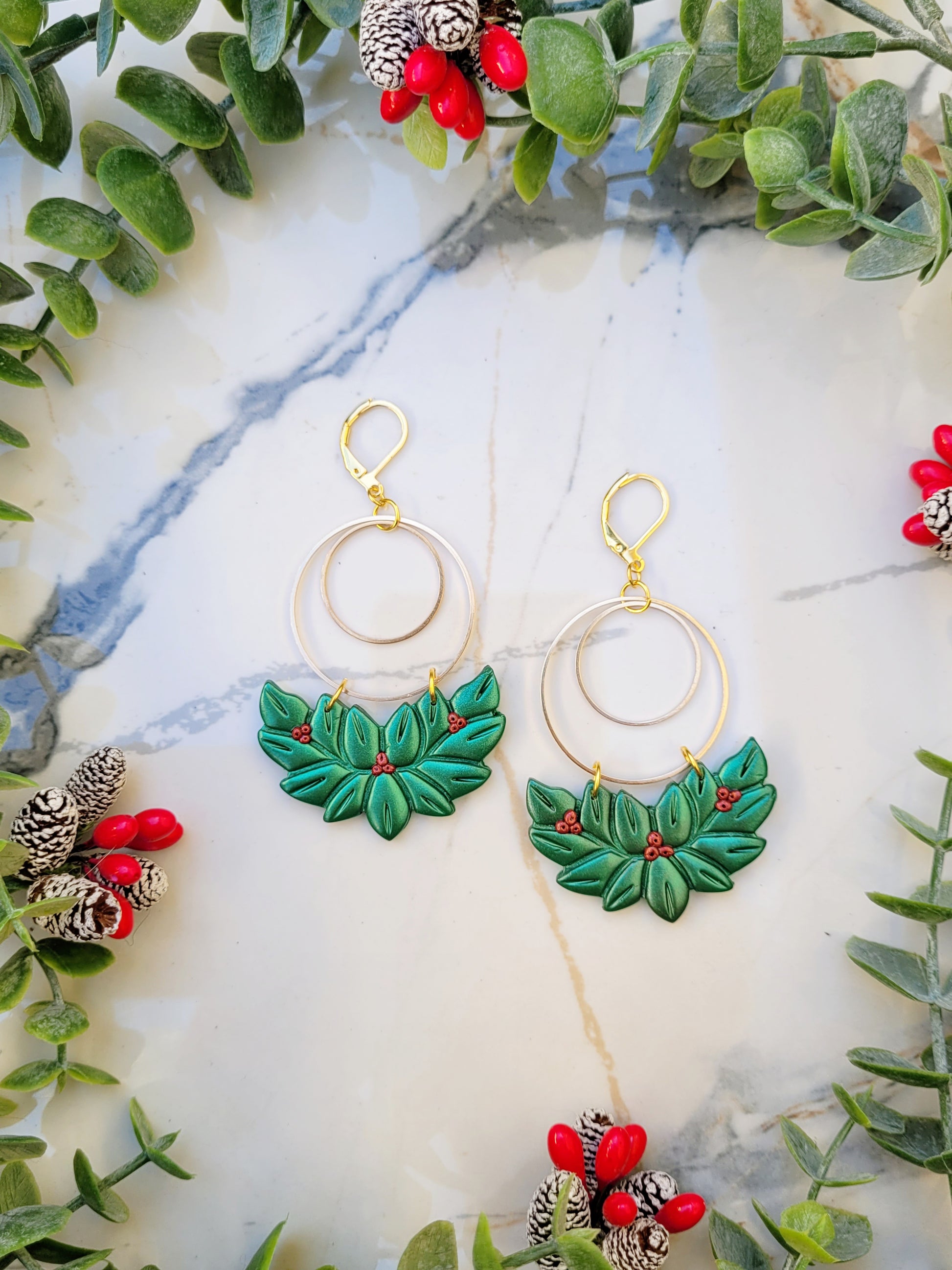 close up of Holly wreath earrings on a marble background surrounded by foliage.
