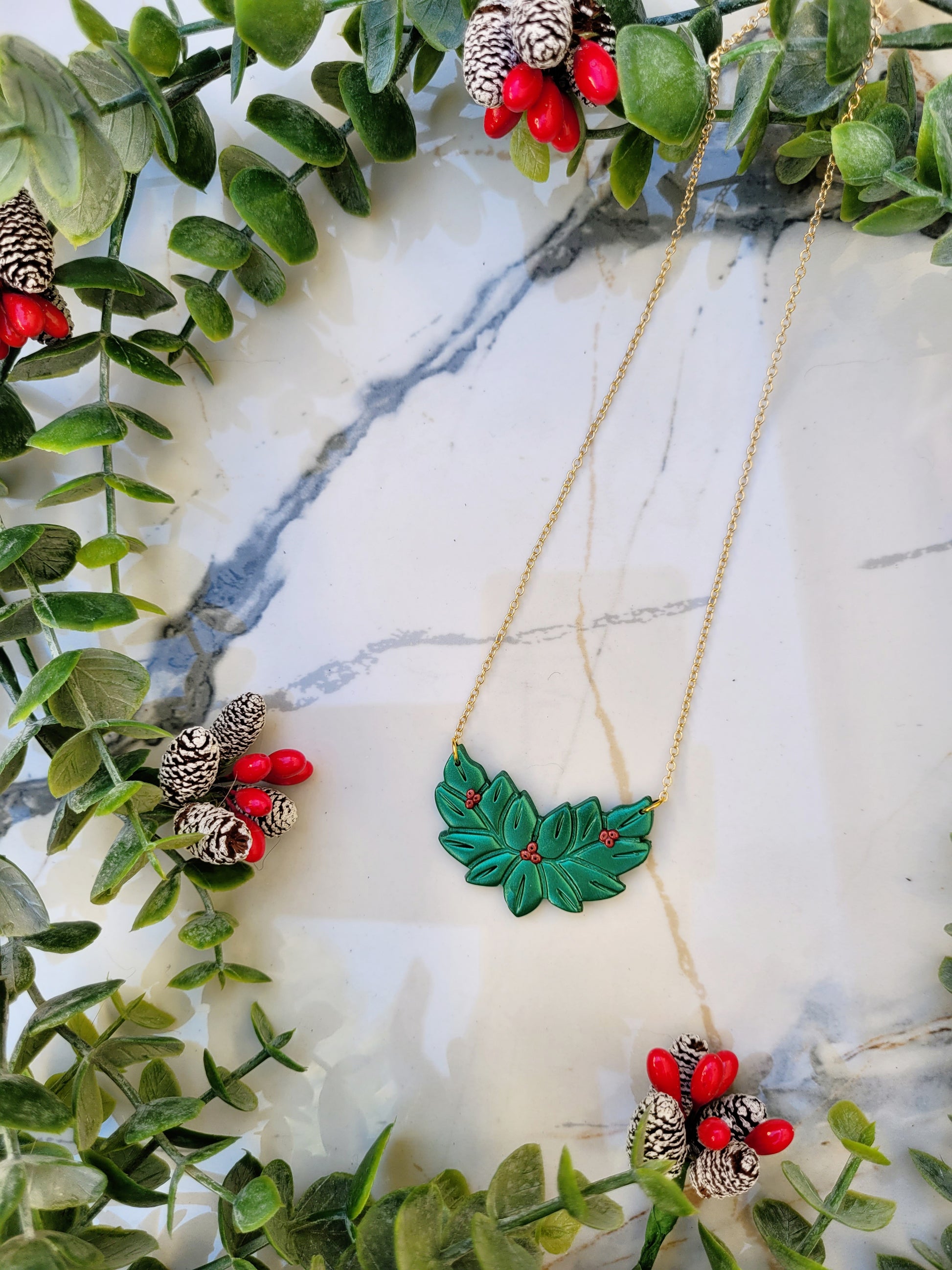 Metallic green and red holly wreath necklace on a marble background surrounded by foliage. 