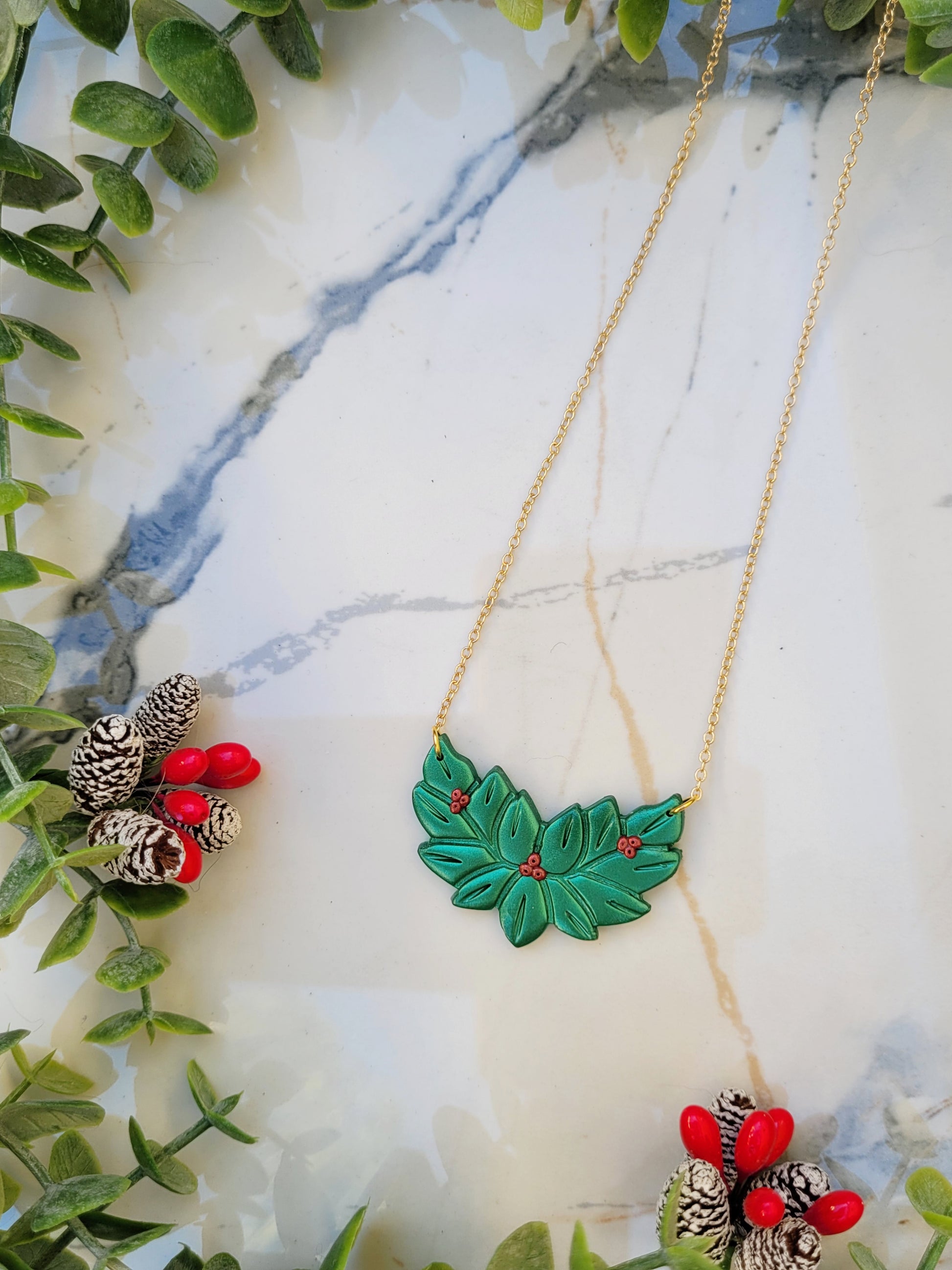close up of Metallic green and red holly wreath necklace on a marble background surrounded by foliage.