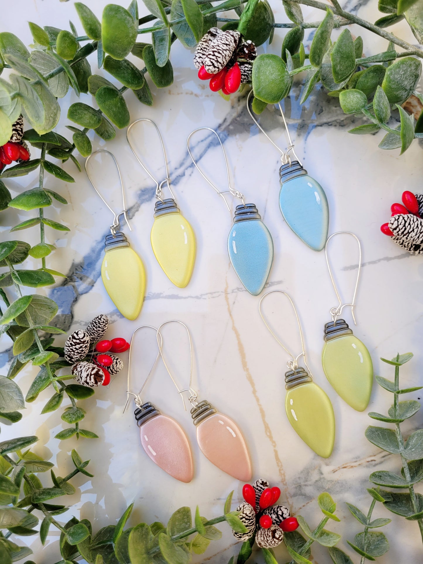 Pink, blue, yellow, and green christmas lightbulb earrings on a marble background surrounded by foliage. 