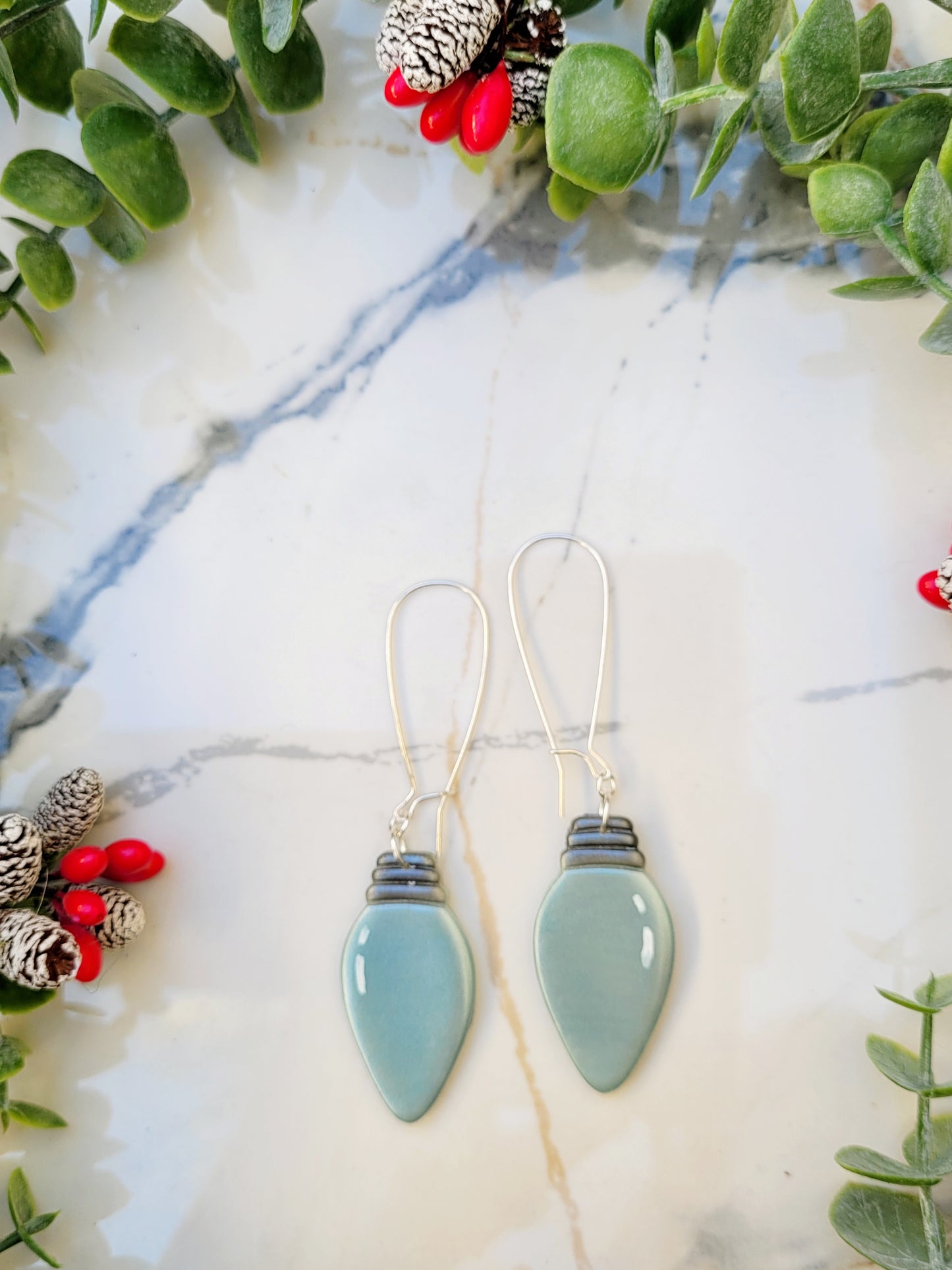 close up of blue christmas lightbulb earrings on a marble background surrounded by foliage.