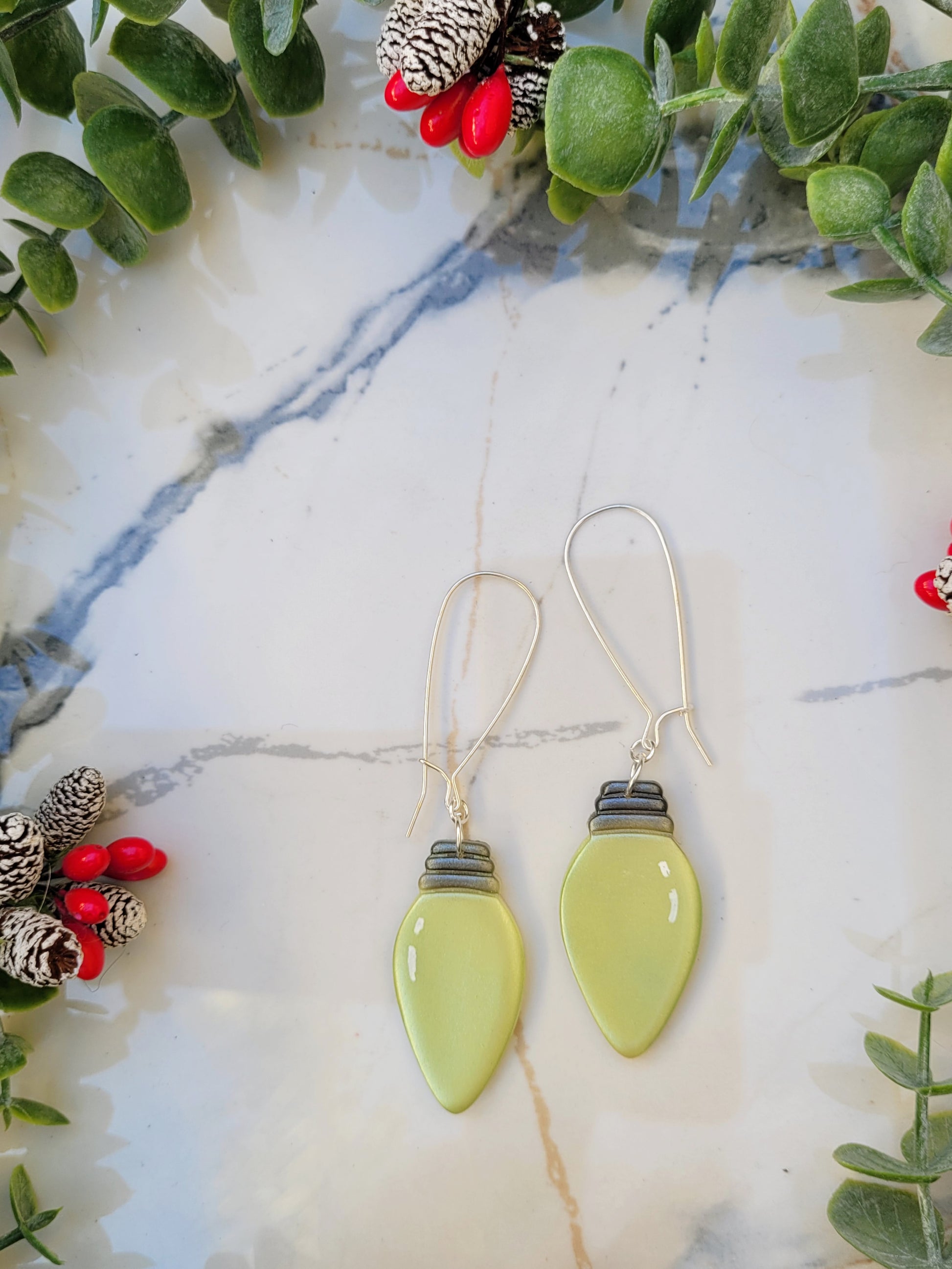 close up of green christmas lightbulb earrings on a marble background surrounded by foliage.