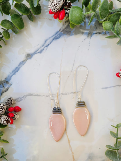 close up of pink christmas lightbulb earrings on a marble background surrounded by foliage.