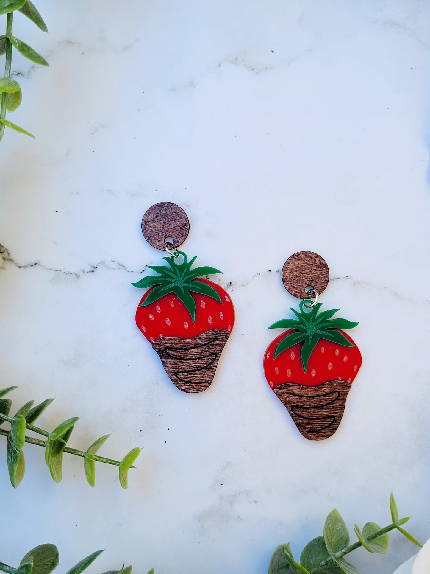 close up of Chocolate covered strawberry earring on white marble background surrounded by foliage.
