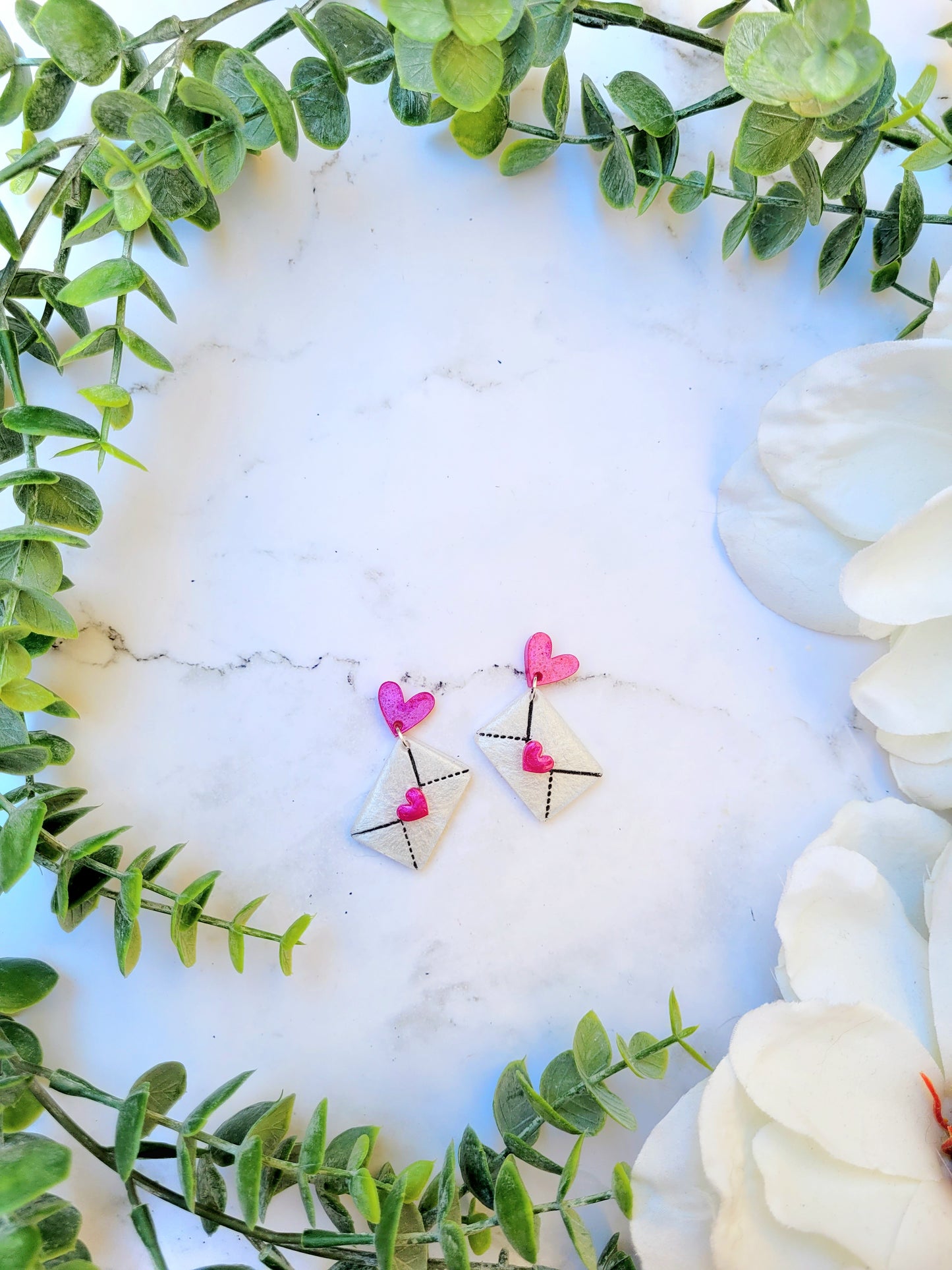 Pink and White Envelope Earrings with Hearts on a white marble background surrounded by foliage. 