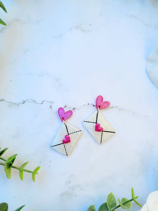 close up of Pink and White Envelope Earrings with Hearts on a white marble background surrounded by foliage. 