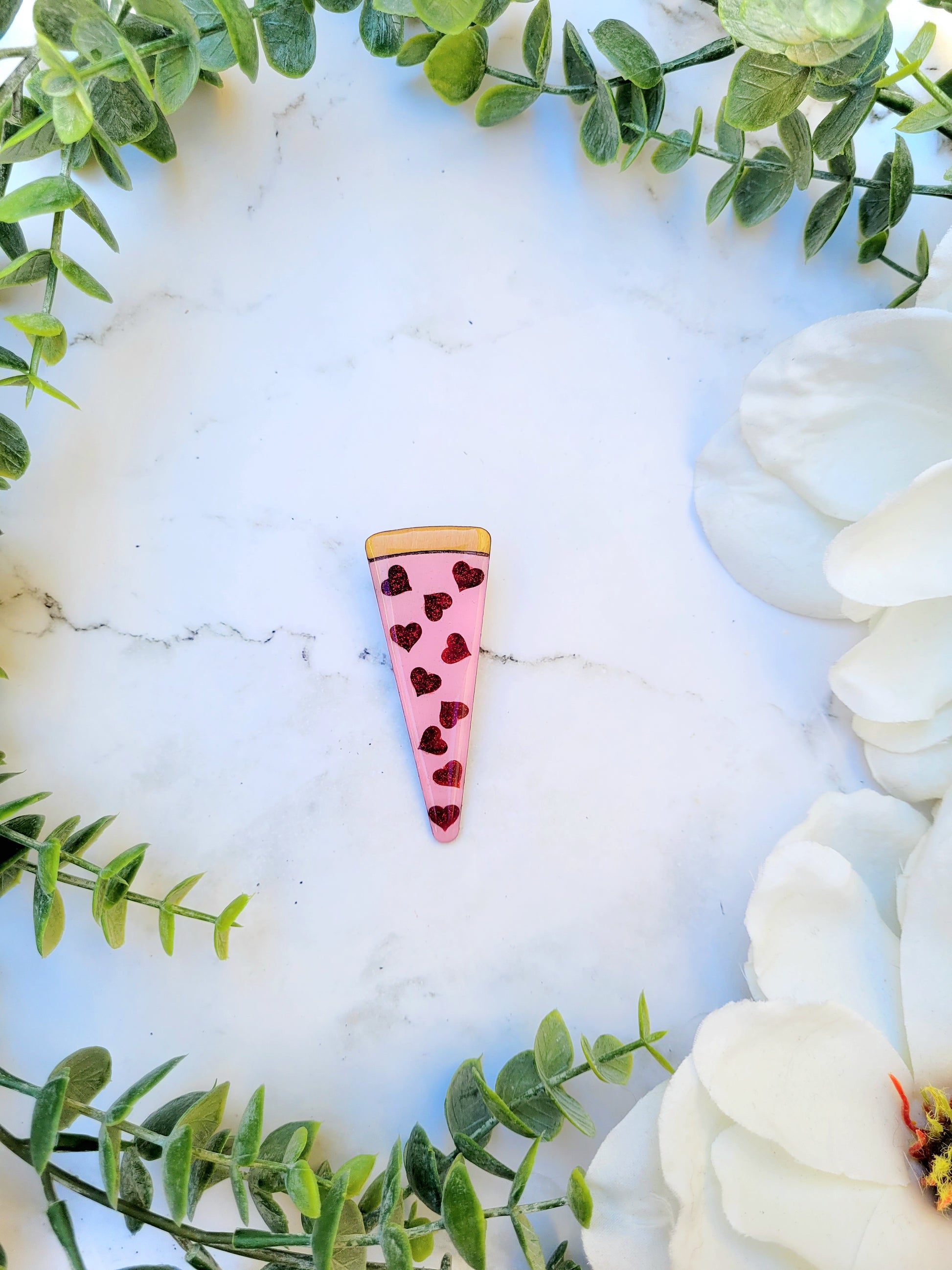Slice of love pizza hair clip on a white marble background surrounded by foliage. 