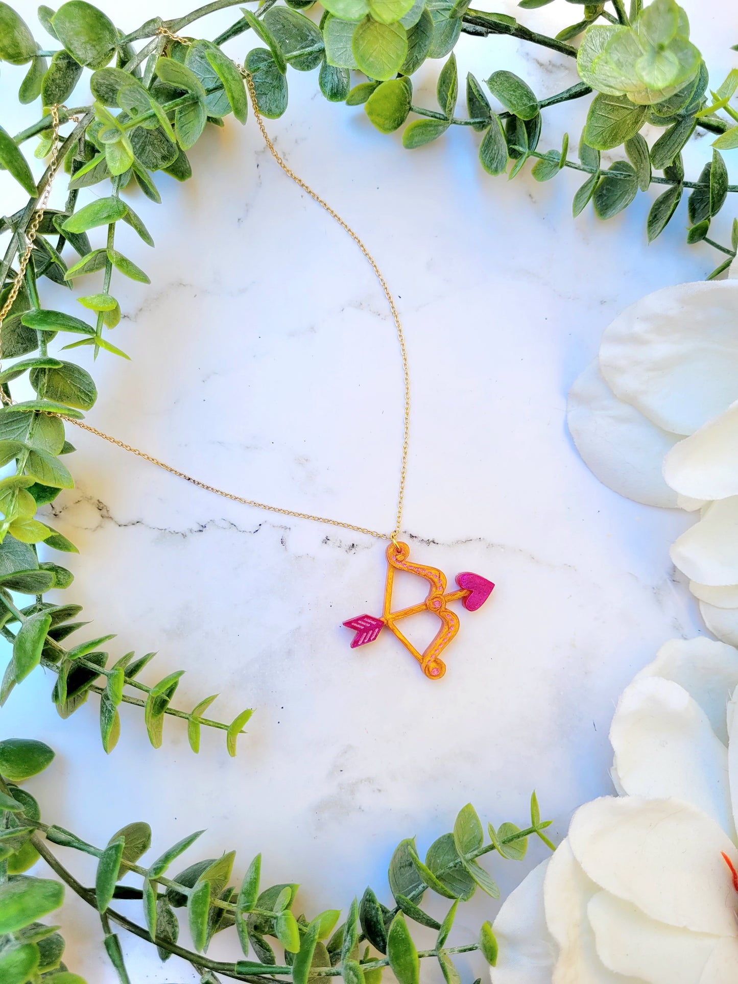 gold and pink cupid's bow necklace on a white marble background surrounded by foliage. 