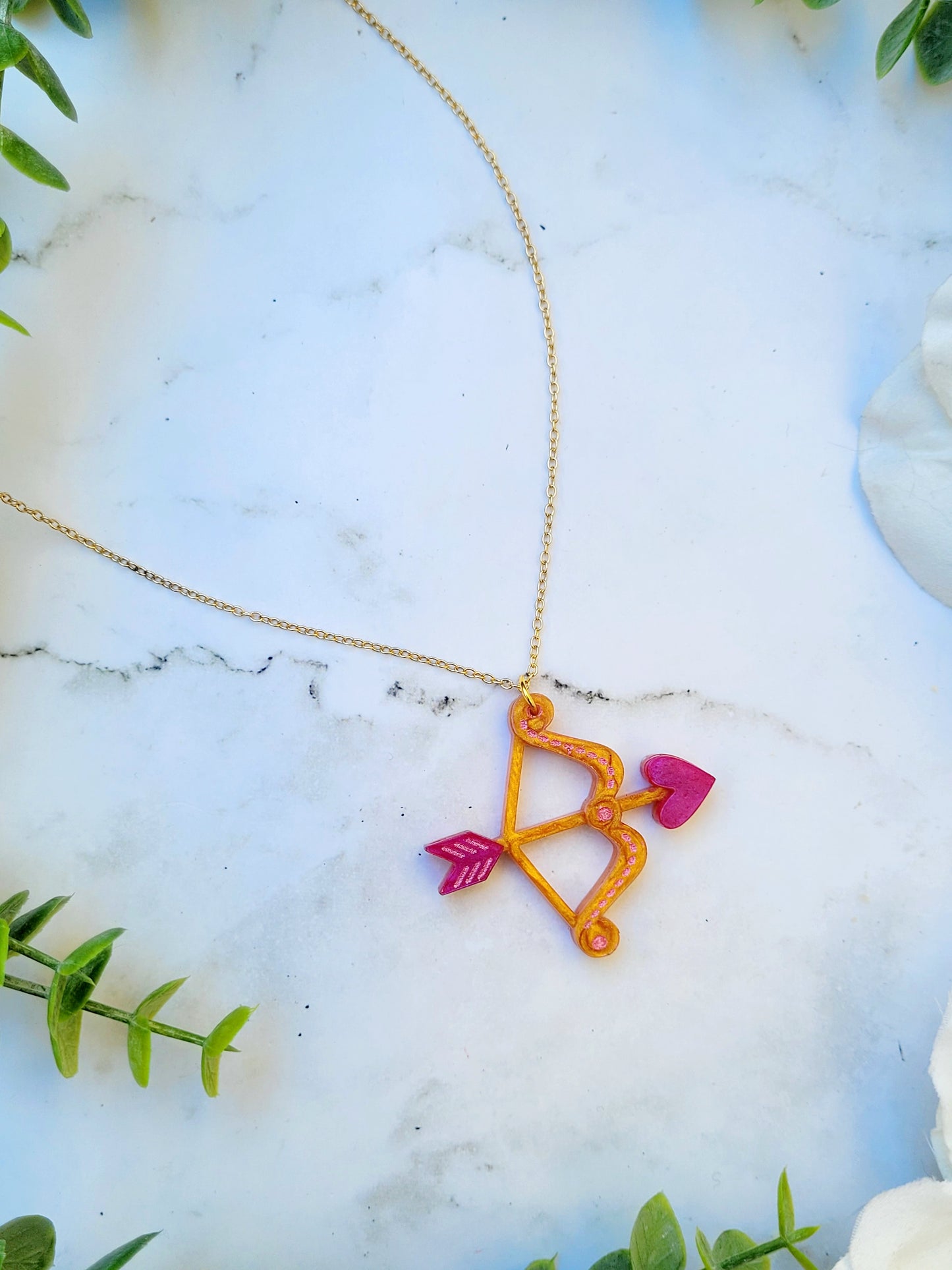 close up of gold and pink cupid's bow necklace on a white marble background surrounded by foliage.