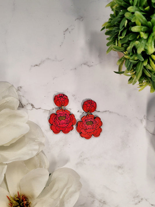 Painted peony earrings on a white marble background surrounded by foliage. 
