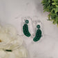 Green pickle earrings on a white marble background surrounded by foliage. 