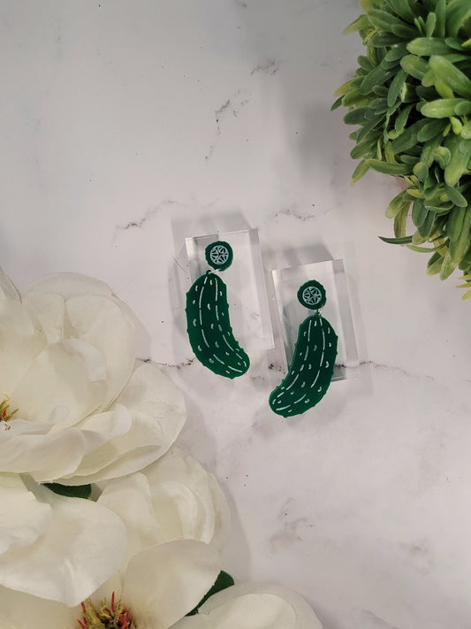 Green pickle earrings on a white marble background surrounded by foliage. 