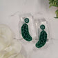 close up of Green pickle earrings on a white marble background surrounded by foliage.