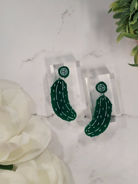 close up of Green pickle earrings on a white marble background surrounded by foliage.