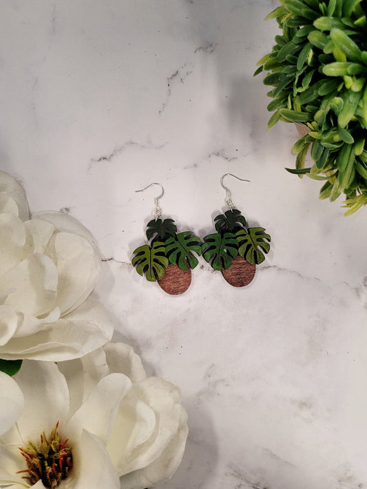 Potted monstera earrings on a marble background surrounded by foliage. 