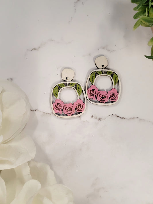 close up of rose garden earrings on a white marble background surrounded by foliage. 