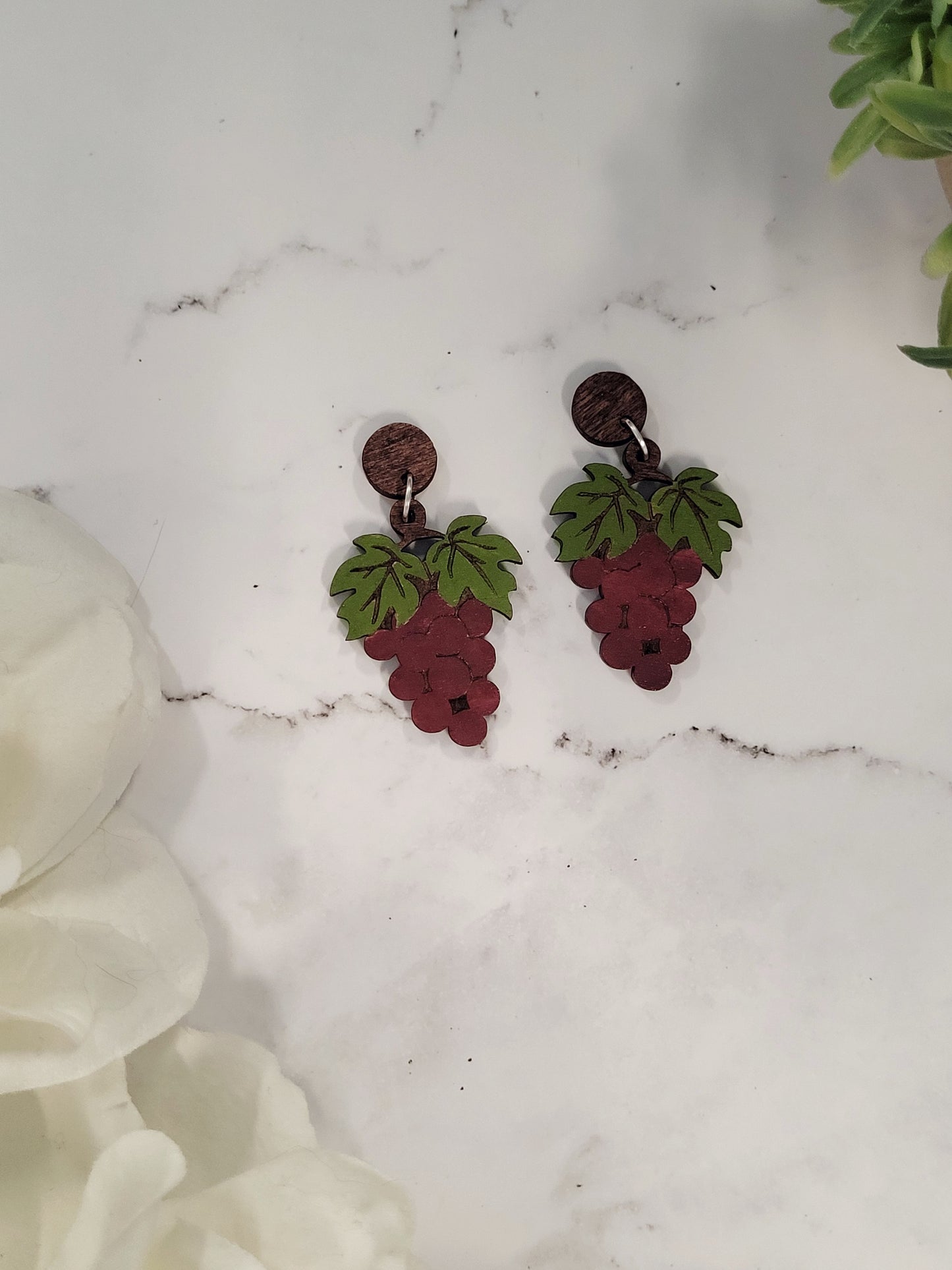 close up of red grape bunch earrings on a marble background surrounded by foliage