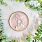faux mother of pearl trinket dish with gold detailing holding a pair of shell earrringson a marble background surrounded by foliage.