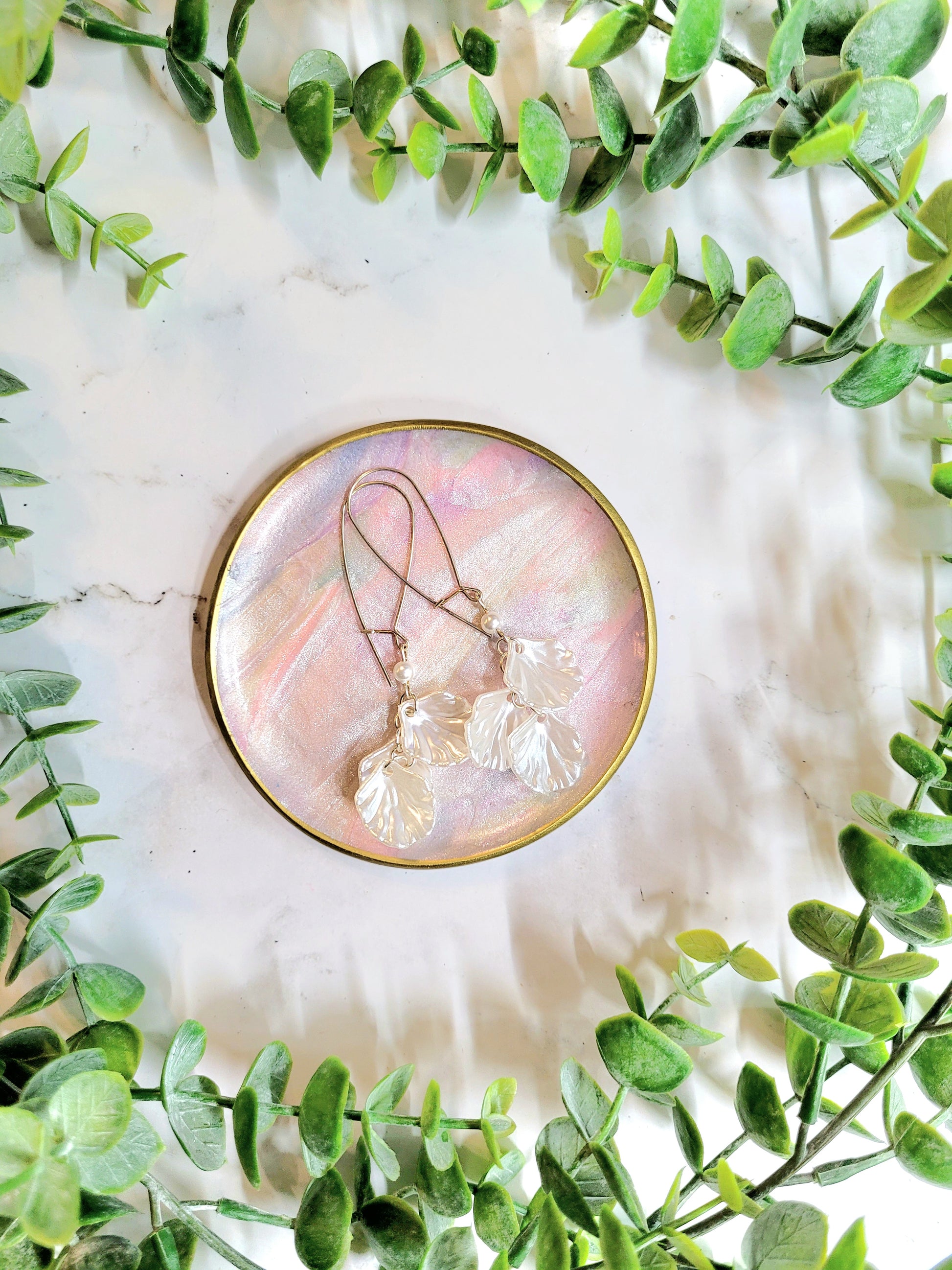 faux mother of pearl trinket dish with gold detailing holding a pair of shell earrringson a marble background surrounded by foliage.