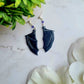 close up of purple bat boy earrings on a marble background surrounded by foliage.