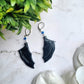 close up of blue bat boy earrings on a marble background surrounded by foliage.