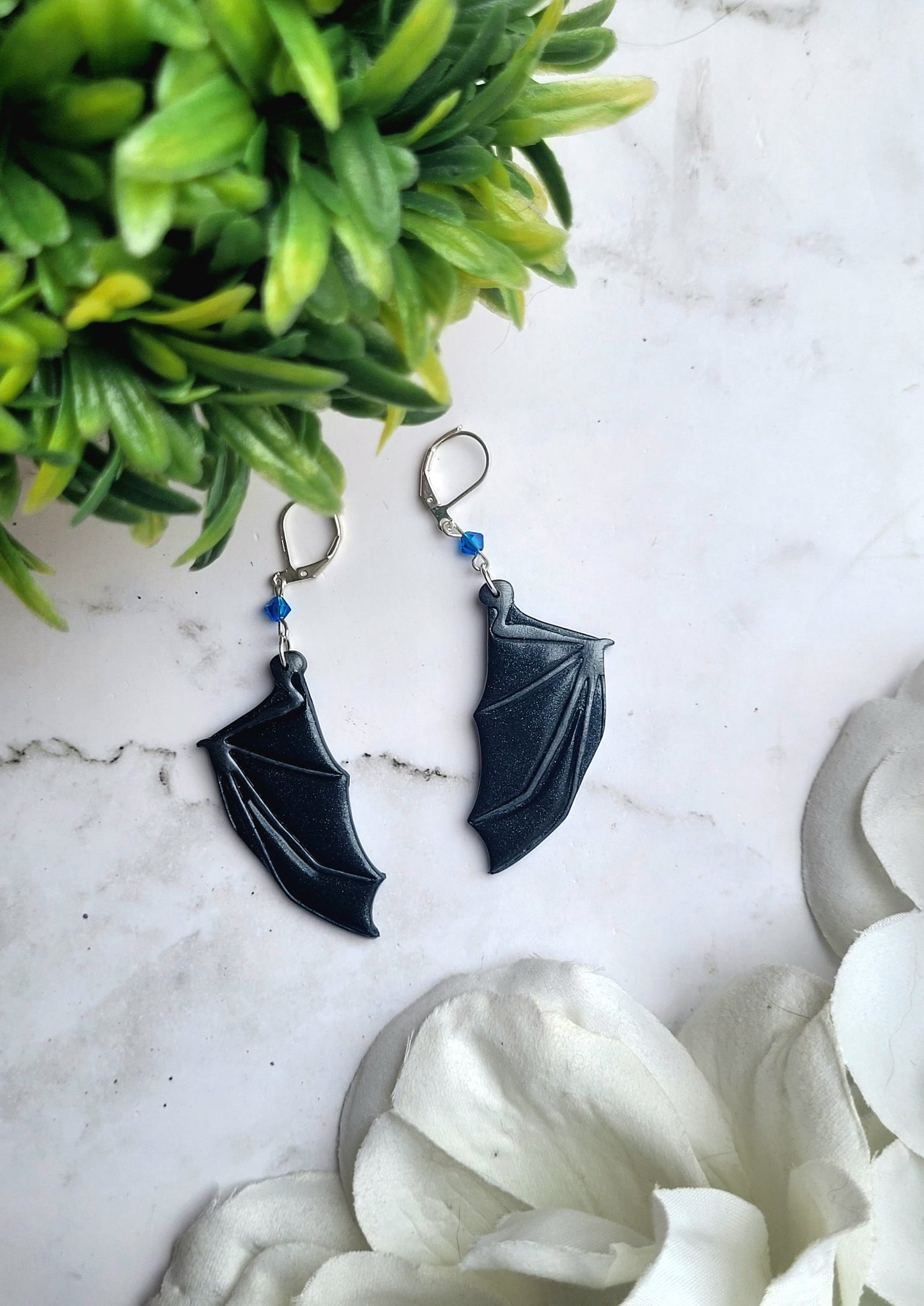 close up of blue bat boy earrings on a marble background surrounded by foliage.