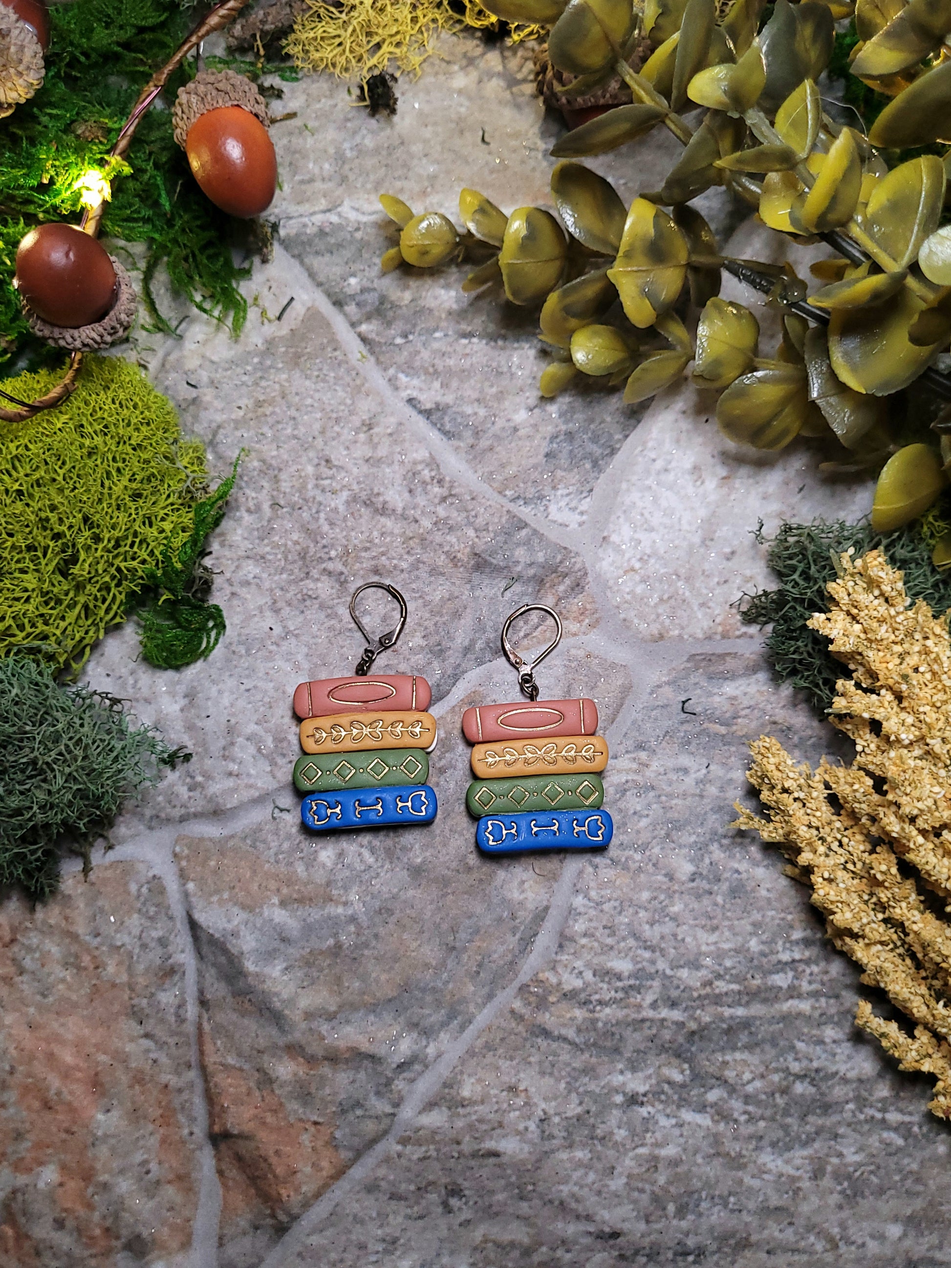 Gilded bookstack earrings on a stone background surrounded by moss and foliage. 