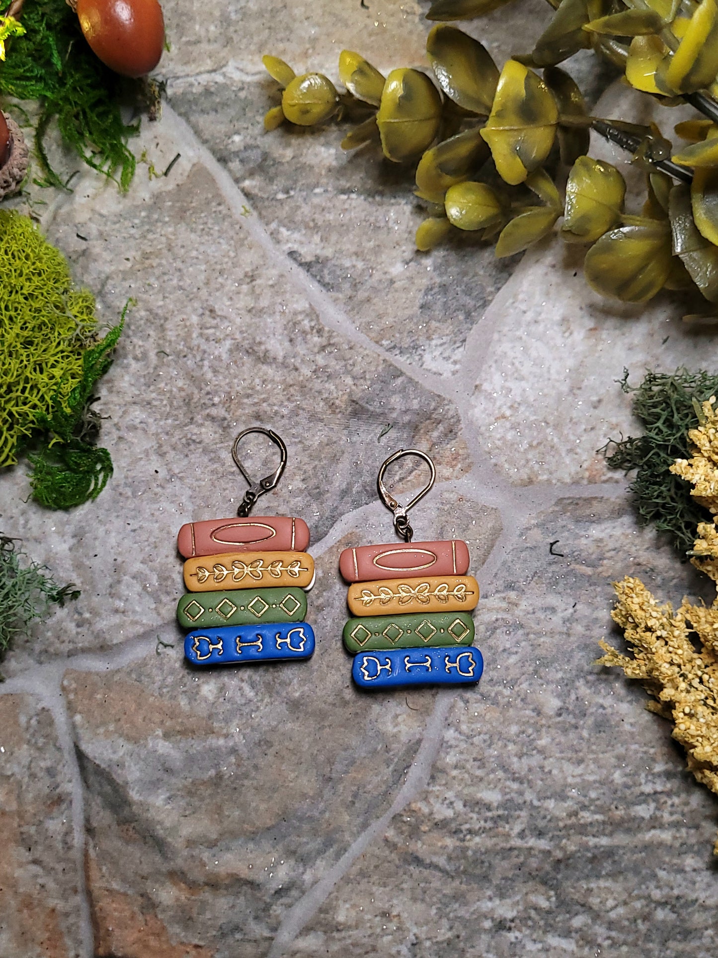 close up of Gilded bookstack earrings on a stone background surrounded by moss and foliage. 