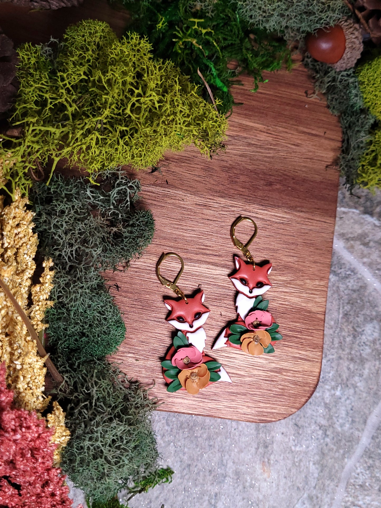 Floral fox earrings on a wood and stone background surrounded by foliage. 