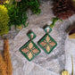 Green diamond shaped earrings with metallic acorns and oak leaves. Sitting on a stone background surrounded by foliage. 