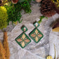 close up of Green diamond shaped earrings with metallic acorns and oak leaves. Sitting on a stone background surrounded by foliage. 