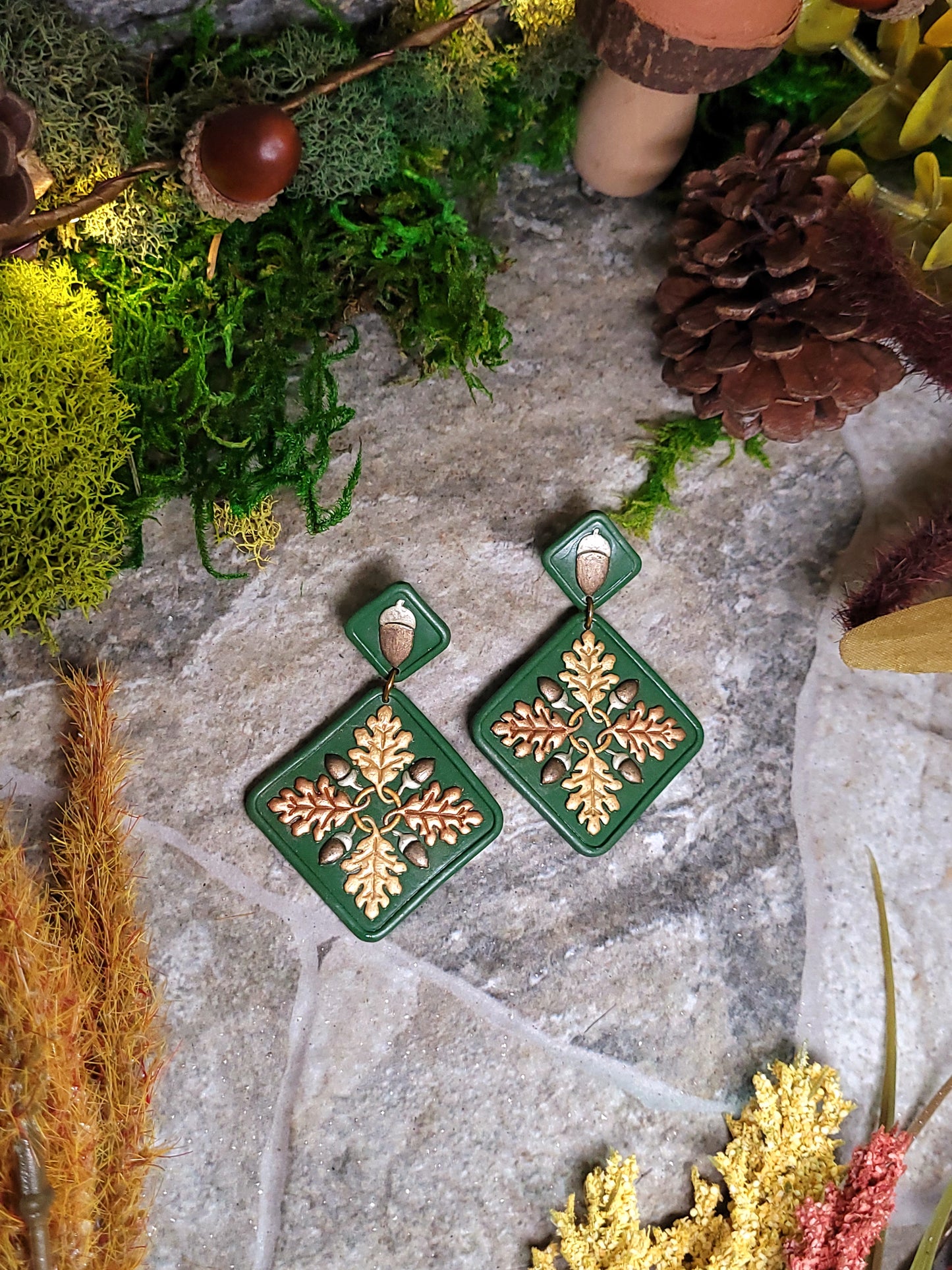 close up of Green diamond shaped earrings with metallic acorns and oak leaves. Sitting on a stone background surrounded by foliage. 