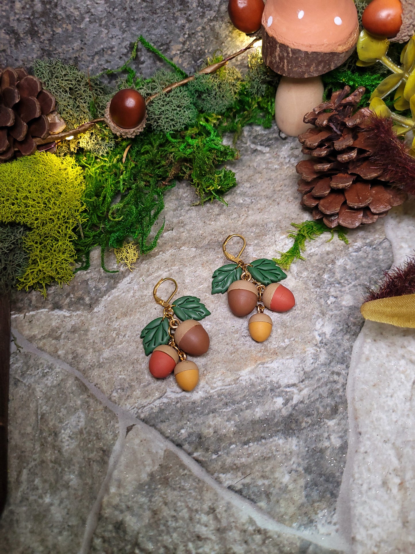 3d acorn shaped earrings with  oak leaves. Sitting on a stone background surrounded by foliage. 