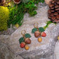 close up of 3d acorn shaped earrings with  oak leaves. Sitting on a stone background surrounded by foliage. 