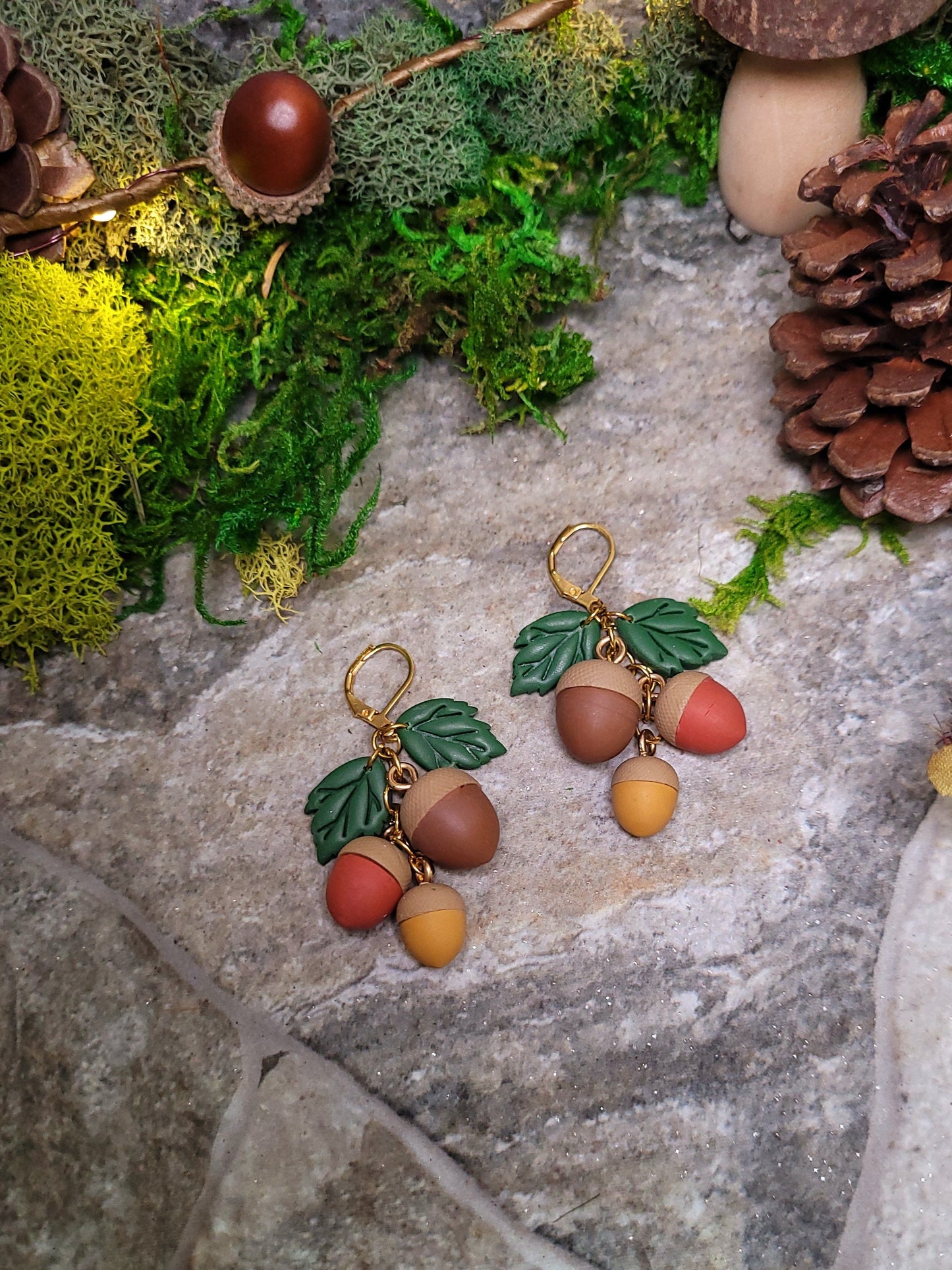 close up of 3d acorn shaped earrings with  oak leaves. Sitting on a stone background surrounded by foliage. 