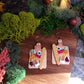 charcuterie board earrings on a wood and stone background surrounded by foliage. 