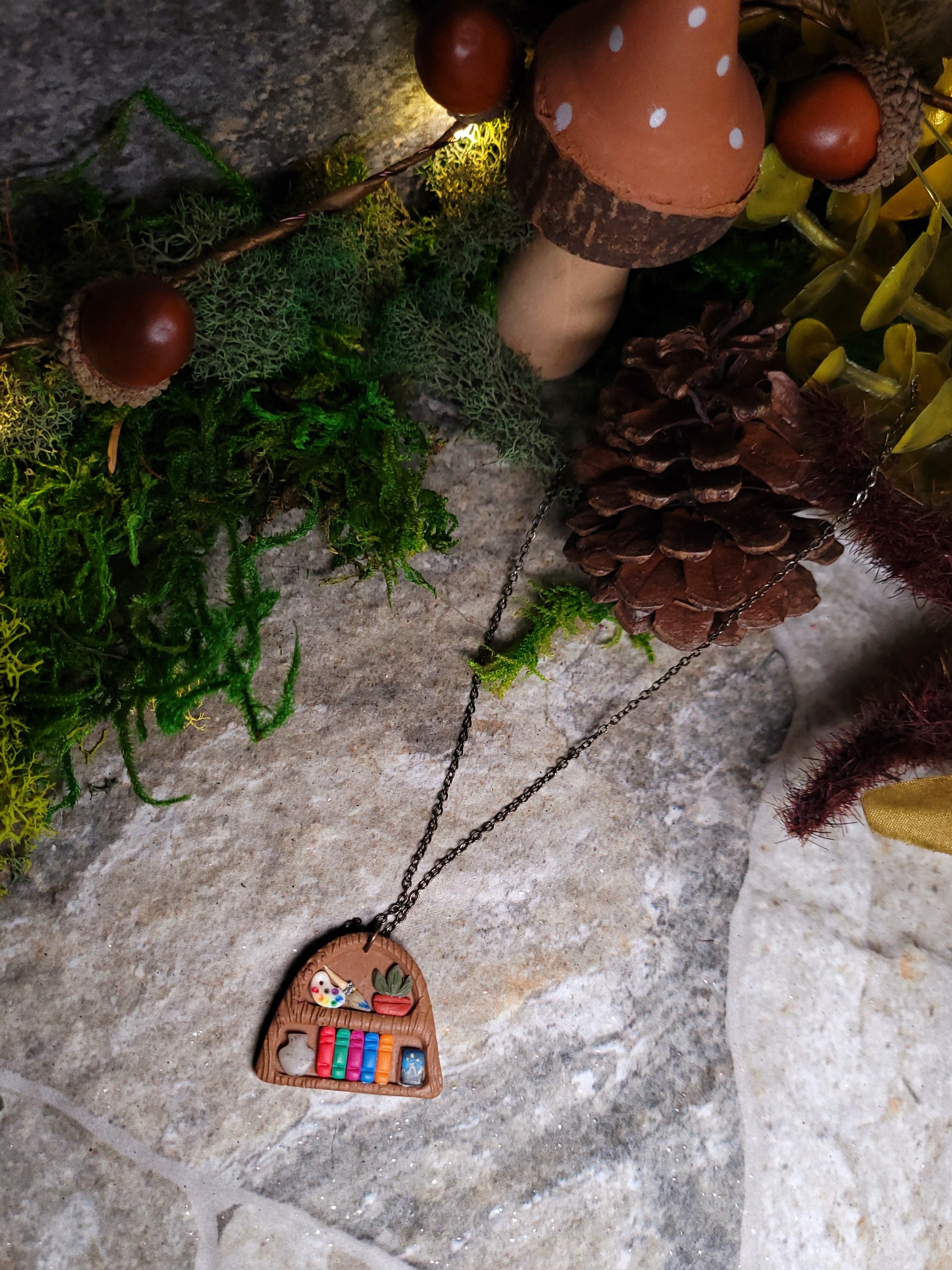 Bookshelf pendant made of polymer clay sits on a stone background surrounded by foliage. 