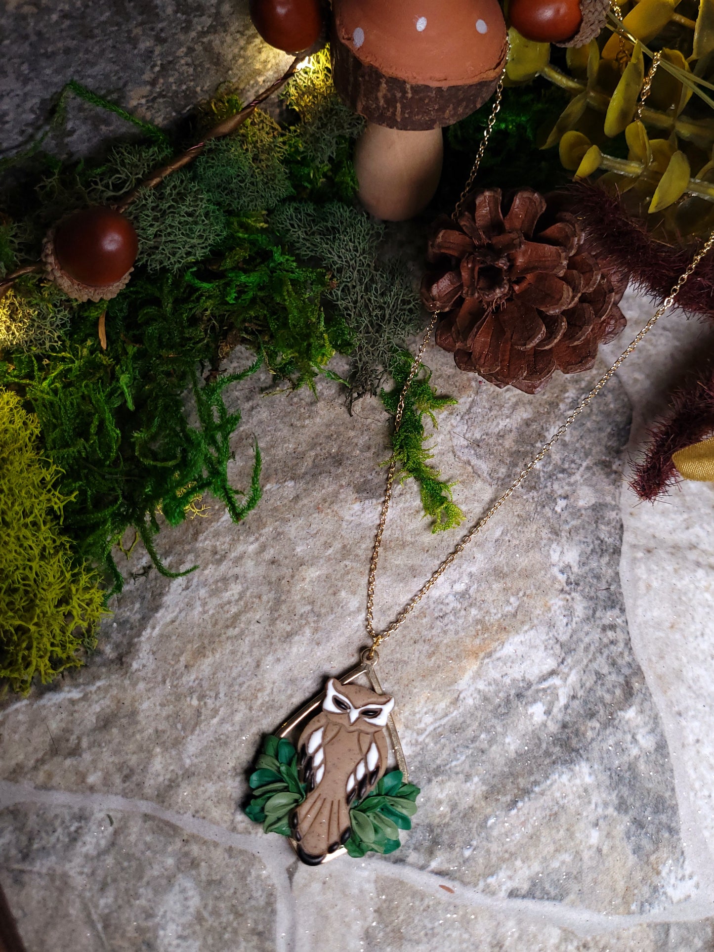 Owl necklace sitting on a stone background surrounded by foliage. 