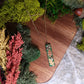  close up of the Green bar shaped pendant with metallic acorns and oak leaves on wood and stone background surrounded by foliage