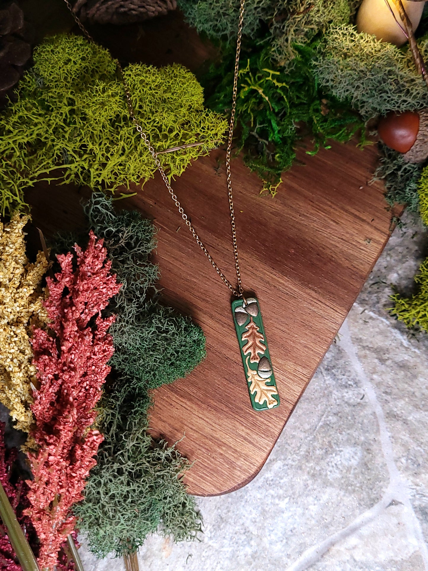  close up of the Green bar shaped pendant with metallic acorns and oak leaves on wood and stone background surrounded by foliage