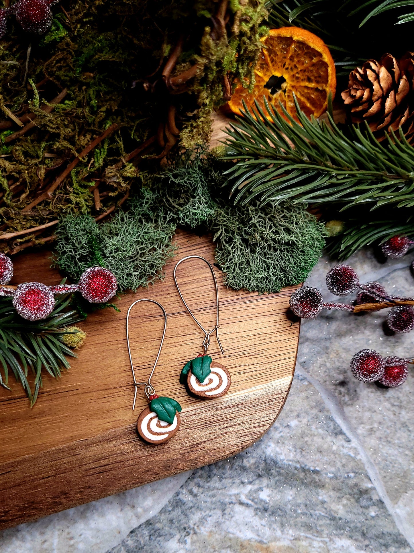 yule log earrings on a wood and stone background surrounded by foliage. 