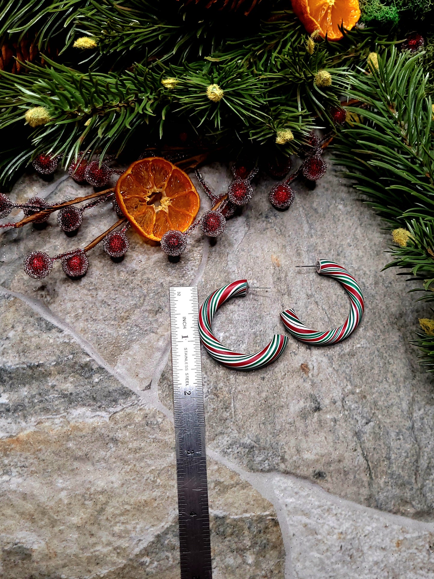 Retro Candy Cane Hoop Earring next to a ruler on a  stone background surrounded by foliage. 