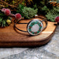 silver holly bracelet on a wood and stone background surrounded by foliage. 