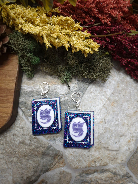 Glitter Dragon Book Earrings on a stone background surrounded by foliage. 