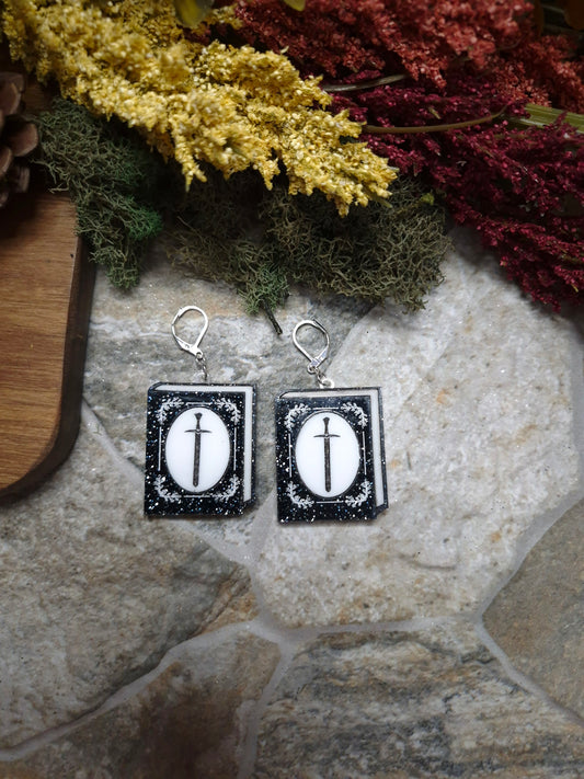 Glitter sword Book Earrings on a stone background surrounded by foliage. 