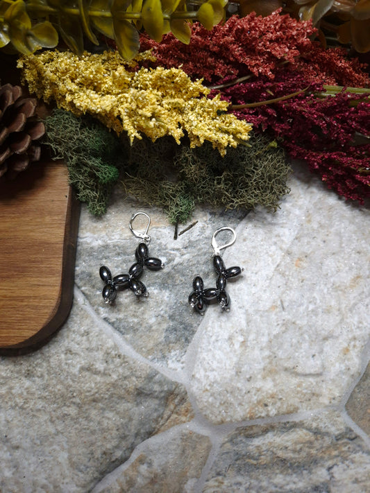 balloon dog earrings on their side on a stone background surrounded by foliage. 