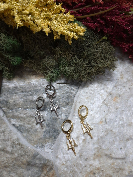Gold and Silver trident earrings on a stone background surrounded by foliage